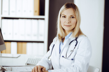 Friendly female doctor sitting in sunny clinic. Portrait of cheerful smiling physician. Medicine and healthcare concept