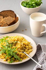 Delicious bulgur porridge with fried onions and herbs in a plate, a glass of milk, arugula in a bowl and black bread