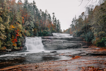 Views of the top two tiers of Triple Falls on the Little River cascading down surrounded by colorful fall foliage on a foggy, misty morning in DuPont State Recreational Forest, North Carolina, USA. 