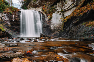 A plunge pool waterfall known as Looking Glass Falls surrounded by logs, stones and colorful fall foliage in Pisgah National Forest, Transylvania County "Land of Waterfalls", North Carolina, USA. 