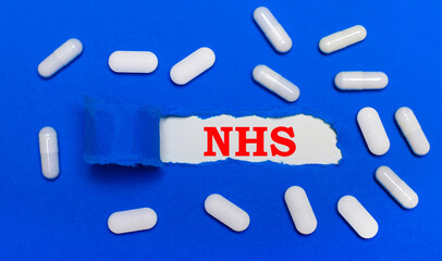 White pills lie on a beautiful blue background. In the center is white paper with the inscription NHS. Medical concept. View from above.