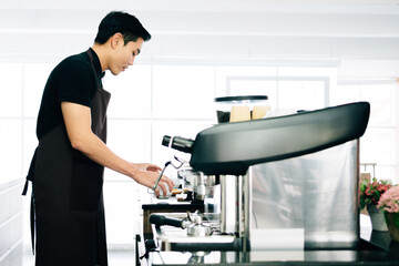 Young Asian male wearing apron preparing coffee cup and standing inside the coffee counter with the coffee machine before making coffee. Business and people's lifestyles during Covid-19 pandemic.