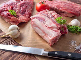 several pieces of fresh raw meat are spread out on parchment. Next to garlic, the herbs, the tomatoes and knife. Side view.