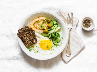 Delicious breakfast, brunch - fried egg, fresh vegetables, hummus, olives, micro greens, grain bread  on a light background, top view