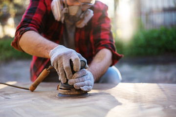 A young man polishes a wooden surface with a grinder in an outdoor respirator, hand made by a carpenter. Craft goods cocept