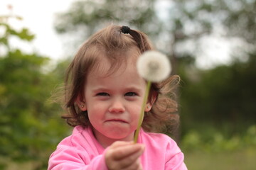 A little girl in a pink T-shirt with long sleeves is blowing on a dandelion she is holding in her hands while standing outdoors.