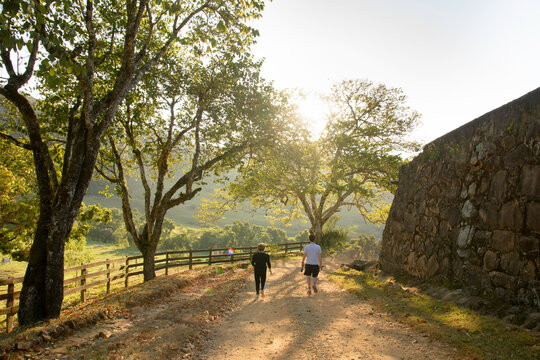 A Man And Woman From The Back Walking On A Dirt Street At Sunset