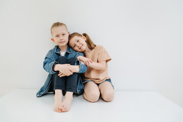 Adorable little siblings, boy and girl, sitting on a table next to each other. Her head is on his shoulder. Over white background.
