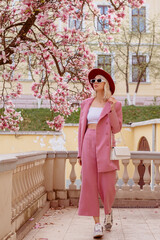 Fashionable woman wearing elegant pink suit, white crop top, sunglasses, burgundy hat, zebra print sneakers, holding white leather bag, walking in street near blooming magnolia tree - obrazy, fototapety, plakaty