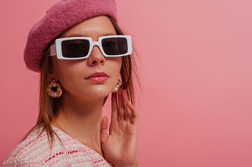 Fashion close up studio portrait of elegant woman wearing classic beret, stylish rectangle white frame sunglasses, trendy pearl earrings, posing on pink background. Copy, empty space for text 