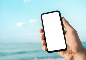 Man holds a smartphone against the background of the sea with a blank screen for mock up.