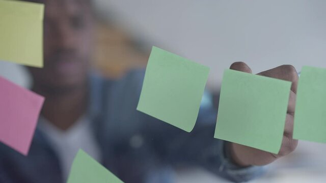 Close-up of sticky notes on glass with African American male hand writing with pen. Blurred young man making notes in home office indoors. Remote working and business ideas