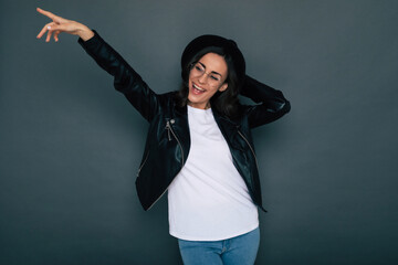 Excited fashionable stylish beautiful young hipster woman in modern black wear and hat while she is posing against the gray wall in the studio and having fun.