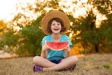 Adorable baby girl in straw hat sit with cross legs on green grass, and eagerly eats juicy...