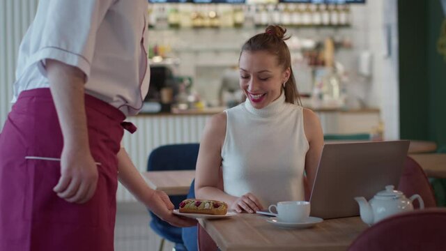 attractive long-haired casually dressed girl works with laptop in cafe, waiter brings her beautiful delicious cake. joys of life, Lockdown cancellation concept. Lifestyle scene in restaurant