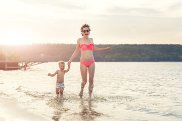 Young adult attractive slim sporty mother enjoy having fun running water by lake or sea sand breach with cute little baby boy against blue sky on summer day. Summertime family vacation concept