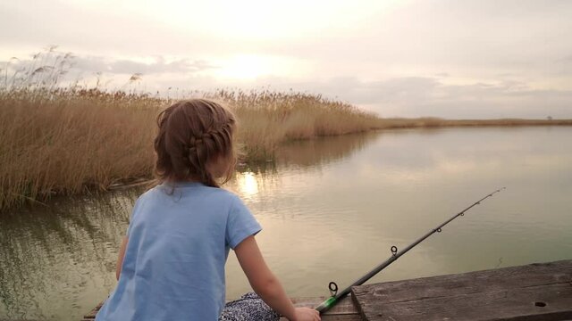 little girl sits on a bridge by the river, takes out a fishing rod without fish