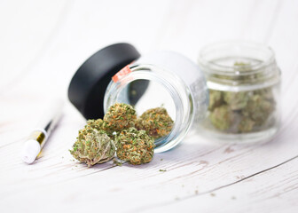 Trimmed cannabis buds up close in glass jars with vape pen on white wood