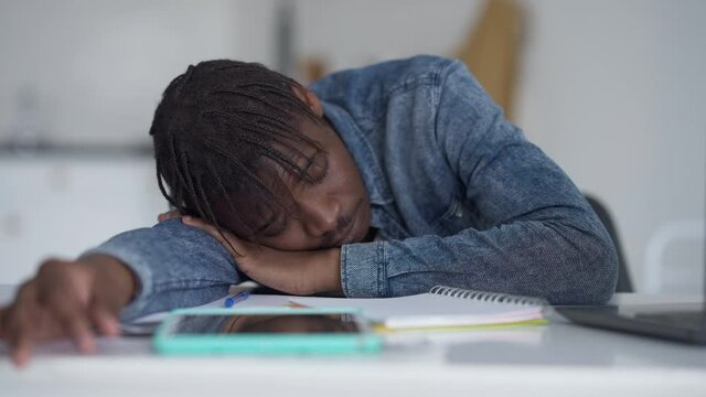 Young tired stressed African American man sleeping on table in home office. Portrait of overworked exhausted handsome guy with closed eyes napping at work time indoors. Burnout concept