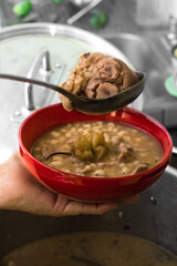Delicious typical Central American dish called pozole served by an adult cook with a metal ladle in a red bowl and a pot of stew made of meat, corn, chili and other succulent ingredients