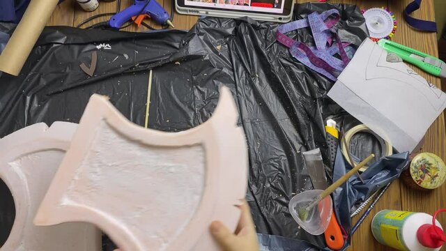 Piece from styrofoam axe in woman's hands. Celtic hatchet made of building materials is covered with soil. Girl takes sandpaper and grinds homemade weapon for cosplayer festival.