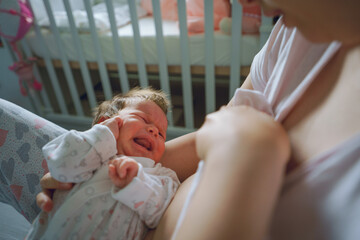 Over the shoulder view on small caucasian newborn baby crying having cramps while her mother is trying to feed her holding in arms breastfeeding problem - obrazy, fototapety, plakaty