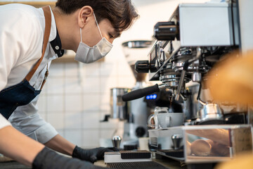 Asian male barista wearing face mask operating coffee machine in cafe.
