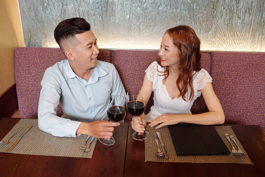 Smiling young boyfriend and gilfriend looking at each other when clinking glasses of wine celebrating first anniversary