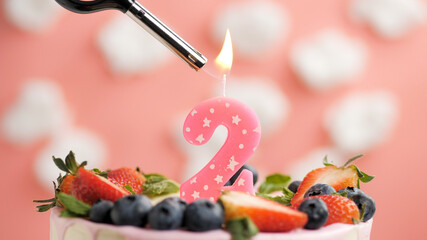 Birthday cake number 2, pink candle on beautiful cake with berries and lighter with fire against background of white clouds and pink sky. Close-up