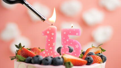 Birthday cake number 15, pink candle on beautiful cake with berries and lighter with fire against background of white clouds and pink sky. Close-up