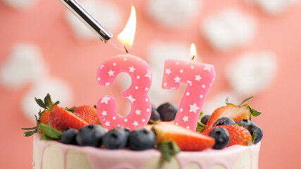 Birthday cake number 37, pink candle on beautiful cake with berries and lighter with fire against background of white clouds and pink sky. Close-up