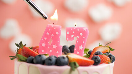 Birthday cake number 47, pink candle on beautiful cake with berries and lighter with fire against background of white clouds and pink sky. Close-up