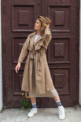 Young smiling millennial woman with wild hair dressed in an autumn coat posing near the door of an...
