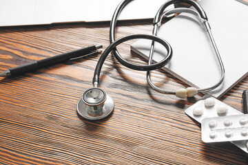 Modern stethoscope, notebook and pills on wooden background, closeup