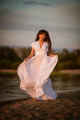Fototapeta na wymiar beautiful, young brunette woman in a white flowing dress on the sandy beach in the evening. full-length portrait in natural light.