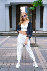 Young beautiful woman of Kakaz nationality poses in the afternoon on the street of the city in a white tracksuit and blue jeans. Confident blonde woman in sunglasses in summer