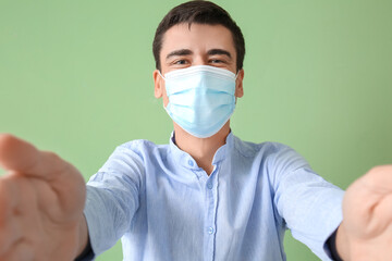 Young man in medical mask opening arms for hug on color background