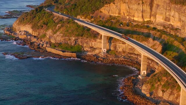 Vehicles Travelling At Sea Cliff Bridge Over Calm Blue Ocean At Sunrise In New South Wales, Australia. - aerial