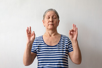 An elderly woman, a gray-haired lady of the 60s in a striped white and blue dress, relax doing meditation gesture with fingers. Yoga concept.