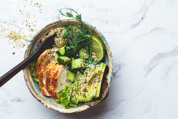Quinoa salad with grilled chicken breast, avocado and cucumber in ceramic bowl on white marble background. Balanced diet concept.