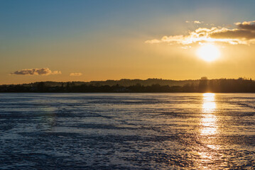 Fototapeta premium Winter landscape with frozen lake at sunrise or sunset. Lake glistening ice reflect a sun.Forest in the background.