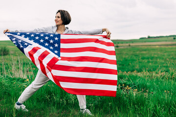 Extremely happy satisfied woman with red lips holding in hands big american flag sincerely rejoicing. Green field on the background. Patriotism