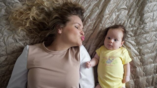 Joyful young mother with curly hair plays with baby daughter in yellow bodysuit kissing girl hand on large bed in apartment room closeup