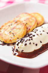 Small cheese pancakes with sour cream and chocolate served in a white plate over wooden table with red plaid tablecloth.