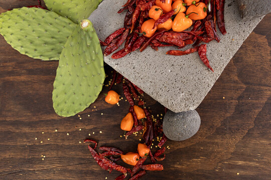 Aerial view of prehispanic foods from Mexico such as metate, chili peppers, prickly cactus, habanero peppers and chile de arbol surrounded by chili seeds on dark wood