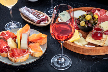 Fototapeta na wymiar Rose wine and Italian antipasti or Spanish tapas in a bar. Blue cheese, prosciutto di Parma ham, jamon, olives and samon sandwiches. Gourmet appetizers on a black background