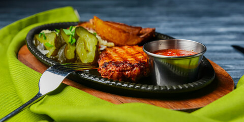 Juicy grilled pork steak with baked potatoes, cut pickles and tomato sauce served in a tray over rustic wooden table.
