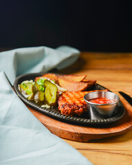 Juicy grilled pork steak with baked potatoes, cut pickles and tomato sauce served in a tray over rustic wooden table.