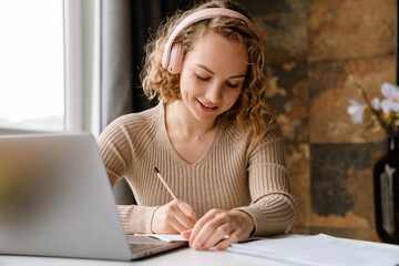 Fototapeta na wymiar Young woman in headphones writing down notes while working with laptop