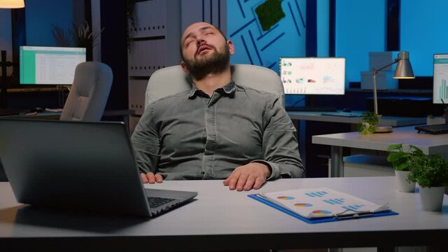 Workaholic businessman slepping on chair in business company office late at night after checking financial statistics on laptop computer. Exhausted manager working overworked at economic project